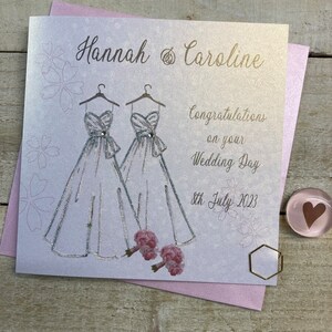 May include: A white card with a silver glitter design of two wedding dresses and a bouquet of pink flowers. The card says "Hannah & Caroline" and "Congratulations on your Wedding Day 8th July 2023".