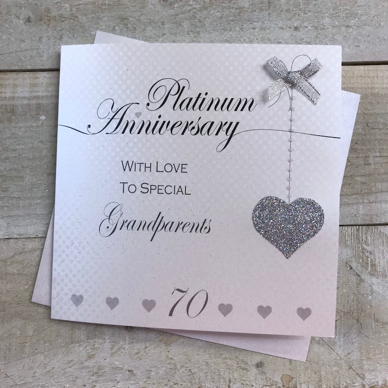 Puede incluir: Una tarjeta blanca con un coraz&oacute;n de purpurina plateada y un lazo plateado. La tarjeta dice "Aniversario de platino" con amor para los abuelos especiales 70.