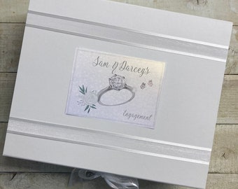 May include: White gift box with a silver ribbon and a personalized label that says "Sam & Darcey's Engagement" with a drawing of a diamond engagement ring.