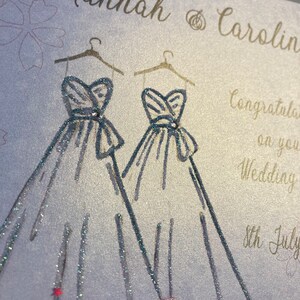 May include: A wedding card with a silver glitter background and two silver glitter wedding dresses drawn on it. The text on the card reads "Hannah & Carolin Congratulations on your Wedding 8th July".