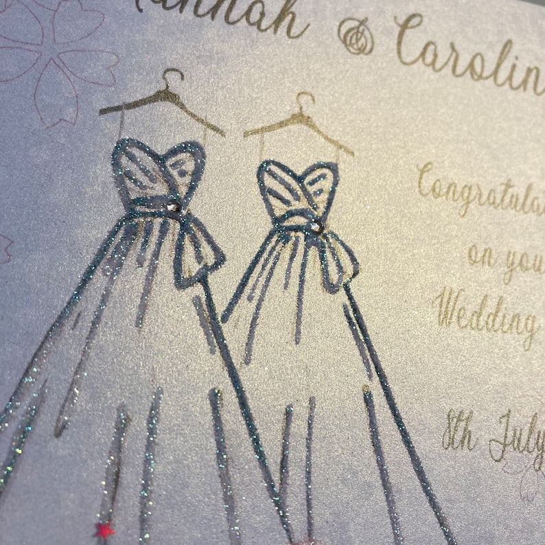 May include: A wedding card with a silver glitter background and two silver glitter wedding dresses drawn on it. The text on the card reads "Hannah & Carolin Congratulations on your Wedding 8th July".