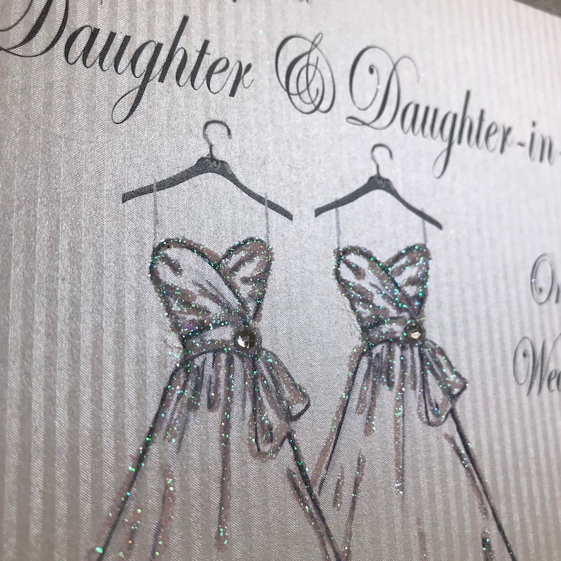 May include: A card with a silver glitter background and the text "To our Daughter & Daughter-in-law On your Wedding Day". Two white dresses with silver glitter are drawn on the card, each hanging on a black hanger.