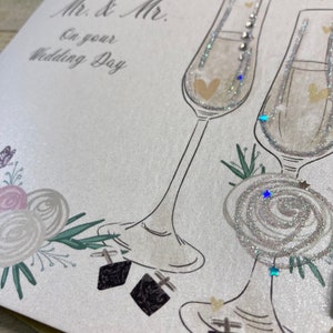 K&ouml;nnte beinhalten: Eine Hochzeitskarte mit silbernem Glitzerhintergrund und einem floralen Design. Die Karte zeigt zwei Champagnergl&auml;ser mit silbernem Glitzerdesign und den Text "Mr. & Mr. On your Wedding Day".