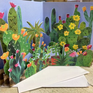 Puede incluir: Una tarjeta pop-up 3D que presenta una escena de jardín colorida y caprichosa con varios cactus, suculentas y flores. La tarjeta está hecha de papel y tiene un diseño en capas, creando un efecto tridimensional.