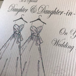 May include: A wedding card with a silver background and two silver dresses drawn on it. The text on the card reads "To a special Daughter & Daughter-in-law On Your Wedding Day".