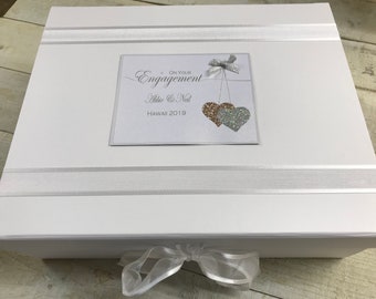 May include: White gift box with a white ribbon and a card that says "On Your Engagement Abbie & Ned Hawaii 2019" with two sparkly hearts hanging from a bow.