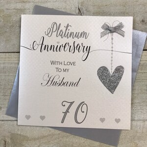 Puede incluir: Una tarjeta blanca con un coraz&oacute;n y un lazo de purpurina plateada. La tarjeta dice "Aniversario de platino" con amor para mi esposo 70.