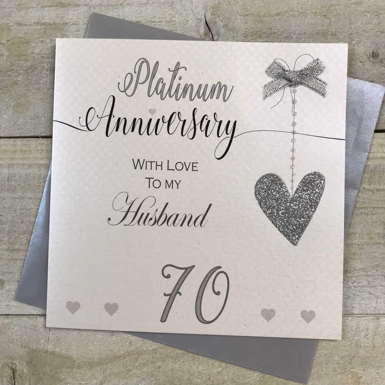 Puede incluir: Una tarjeta blanca con un coraz&oacute;n y un lazo de purpurina plateada. La tarjeta dice "Aniversario de platino" con amor para mi esposo 70.
