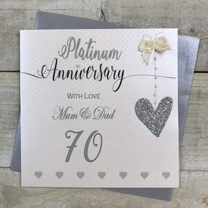 Puede incluir: Una tarjeta blanca con un coraz&oacute;n de purpurina plateada y un lazo plateado. La tarjeta dice "Aniversario de Platino" con el n&uacute;mero "70" debajo. La tarjeta tambi&eacute;n dice "Con amor Mam&aacute; y Pap&aacute;".