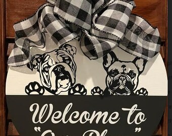 May include: A round wooden sign with a black and white checkered bow. The sign has a white background with two black and white dog silhouettes peeking over the top. The bottom half of the sign is black with the text "Welcome to "Our Place"" in white.