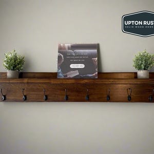 May include: A dark brown wooden shelf with five black metal hooks. The shelf has a small ledge on the top and is mounted on a light gray wall. There is a framed print on the shelf with the text "Funny, I've been in the right place all along. I just didn't know they were looking for me. This wasn't my job.  -  PRISCILLA QUEEN", and two small potted plants on either side of the print. The shelf is labeled "UPTON RUSTICS SOLID WOOD HOME GOODS".