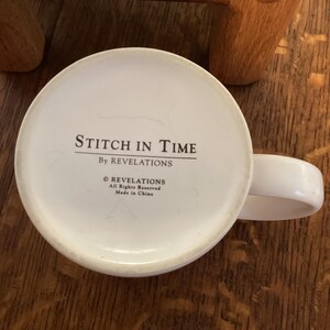 May include: White ceramic mug with a curved handle. The bottom of the mug features the text "Stitch in Time" and "By Revelations". Additional text includes "&copy; Revelations All Rights Reserved Made in China".