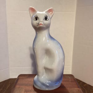 May include: A ceramic cat figurine in a seated pose. The cat is primarily white with blue accents on the back and tail. It has pink-lined ears, black eyes, and a small pink nose and mouth. The figurine is on a wooden surface.