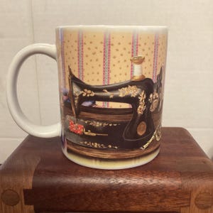 May include: White ceramic mug featuring a black antique sewing machine design with gold accents. The mug has a white handle and a background with a repeating pattern of pink and yellow stripes. A spool of thread is also visible.