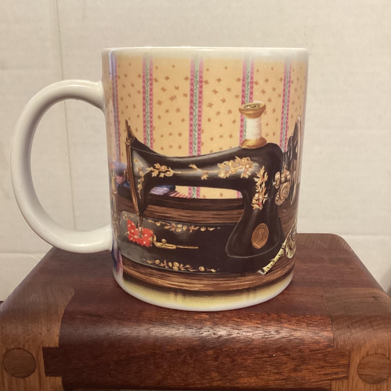 May include: White ceramic mug featuring a black antique sewing machine design with gold accents. The mug has a white handle and a background with a repeating pattern of pink and yellow stripes. A spool of thread is also visible.