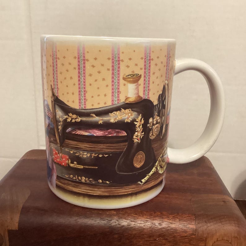 May include: A white ceramic mug featuring a vintage sewing machine design. The mug has a handle and a decorative background with pink and yellow stripes and floral accents. The sewing machine is black with gold detailing.
