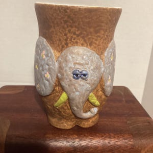 May include: A ceramic vase with a brown textured exterior and an elephant design. The elephant has a gray head, blue eyes, and yellow tusks. Gray ears with pink flowers are on either side of the elephant's head. The vase is on a wooden surface.