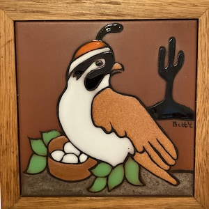 May include: A framed ceramic tile featuring a stylized bird with white, brown, and black plumage, wearing an orange cap. The bird is perched near a nest with eggs, with a black cactus silhouette in the background. The tile is framed in a light brown wooden frame.