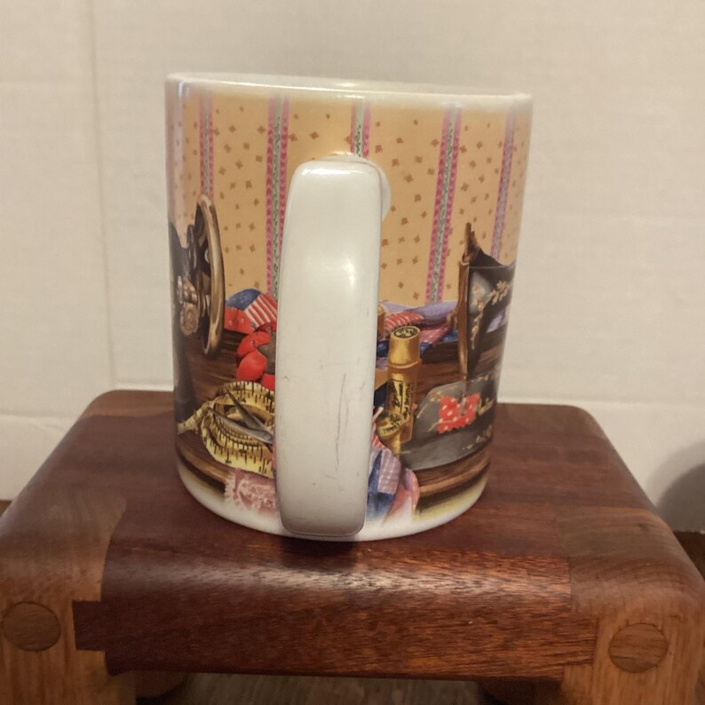 May include: A ceramic coffee mug with a white handle. The mug features a design of sewing tools, including a sewing machine, measuring tape, and spools of thread. The background has a beige and pink pattern.