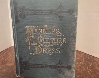 Op de afbeelding: Antiek boek getiteld "Manners and Culture and Dress" met gouden letters op een teal-kleurige omslag. De omslag heeft sierlijke decoratieve elementen in zwart. Het boek staat rechtop op een houten oppervlak.