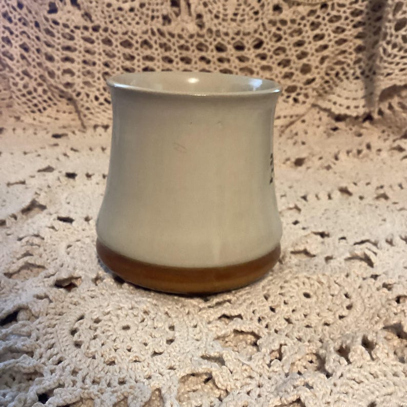 Pu&ograve; includere: Una tazza in ceramica con un corpo beige chiaro e una base marrone. La tazza ha una forma leggermente svasata e una finitura liscia e lucida. La tazza &egrave; alta circa 10 cm.