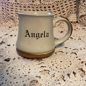 Pu&ograve; includere: Tazza in ceramica color crema con base e manico marroni. Il nome "Angela" &egrave; stampato in lettere gotiche nere sul davanti. La tazza &egrave; appoggiata su una superficie all'uncinetto color crema.