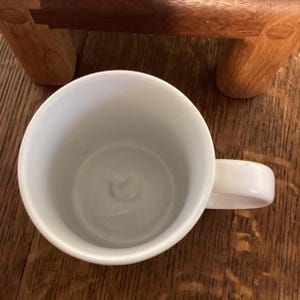 May include: A white ceramic mug with a curved handle. The mug is empty and sits on a wooden surface. A portion of a wooden object is visible in the background.