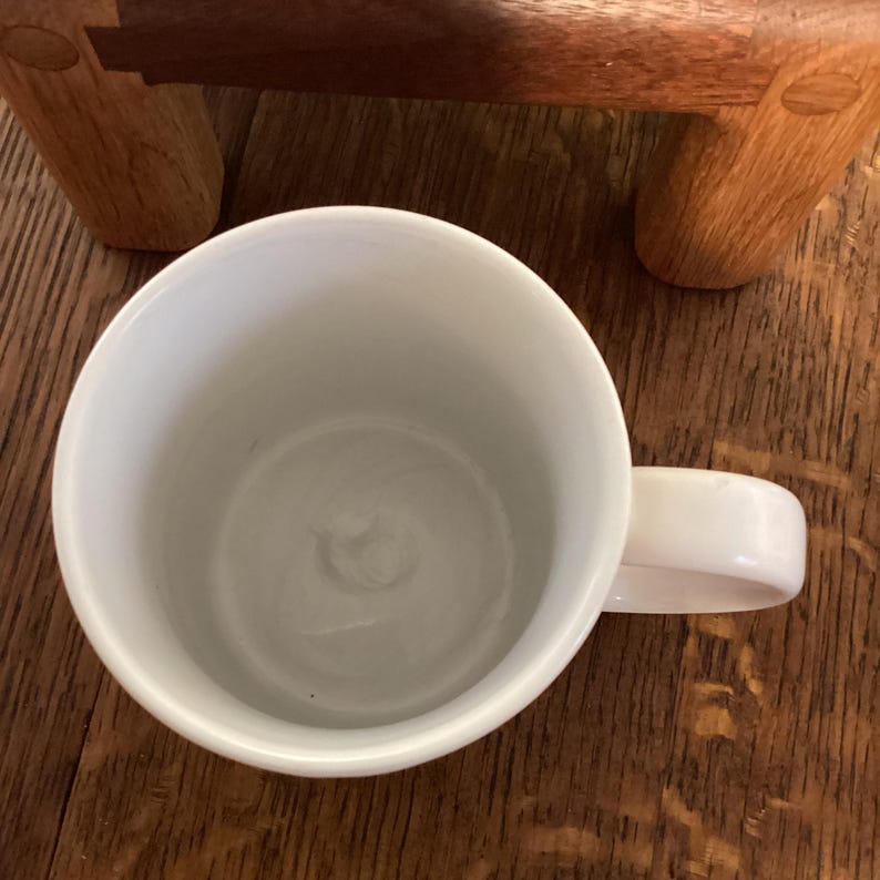 May include: A white ceramic mug with a curved handle. The mug is empty and sits on a wooden surface. A portion of a wooden object is visible in the background.