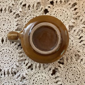 Pu&ograve; includere: Una tazza in ceramica marrone con manico. La tazza ha una forma circolare con un interno pi&ugrave; chiaro e un esterno marrone scuro. Il manico &egrave; piccolo e arrotondato. La tazza &egrave; appoggiata su una superficie all'uncinetto color crema.
