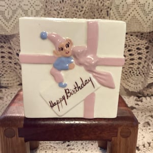 May include: A white ceramic gift box with a pink ribbon design and a small elf figurine. The box has a tag that reads "Happy Birthday" in black script. The elf wears a pink hat and blue outfit.