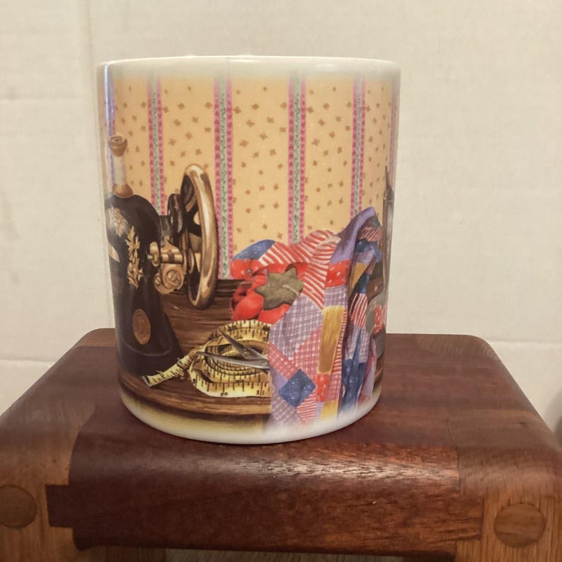 May include: A white ceramic mug featuring a vintage sewing machine, measuring tape, and a patchwork quilt design. The background has a yellow dotted pattern with vertical stripes. The mug is displayed on a wooden stool.