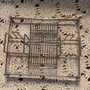 May include: A silver-colored metal napkin holder with a rectangular base and a central section for napkins. The design includes a section for holding utensils. The holder is made of thin metal rods and has a simple, functional design.