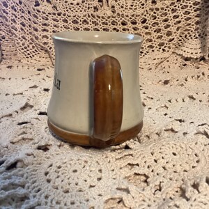 Pu&ograve; includere: Una tazza in ceramica con corpo beige chiaro e manico marrone. Anche la base e il bordo della tazza sono marroni. La parola "la" &egrave; visibile sul lato della tazza. La tazza &egrave; appoggiata su una superficie all'uncinetto.