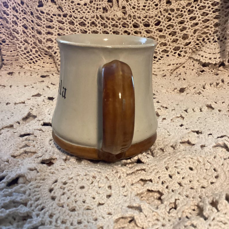 Pu&ograve; includere: Una tazza in ceramica con corpo beige chiaro e manico marrone. Anche la base e il bordo della tazza sono marroni. La parola "la" &egrave; visibile sul lato della tazza. La tazza &egrave; appoggiata su una superficie all'uncinetto.