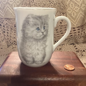 May include: A white ceramic mug featuring a detailed illustration of a fluffy grey kitten with bright blue eyes. The mug has a gold-toned rim and a curved handle. A copper-colored US penny is placed next to the mug for scale reference.