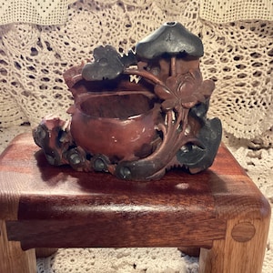 May include: A carved wooden decorative piece with a dark reddish-brown finish. The piece features a central bowl-like structure, a small house-like element, and intricate floral and leaf carvings. The piece is displayed on a small wooden stand.