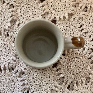 Pu&ograve; includere: Una tazza da caff&egrave; in ceramica color crema con un manico marrone. L'interno della tazza &egrave; di colore verde chiaro. La tazza &egrave; vista dall'alto.