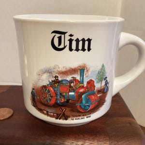 May include: White ceramic mug with a handle. The mug features the name "Tim" in dark blue script. Below the name is a colorful illustration of a steam roller with the text "STEAM ROLLER by AVELING 1893".