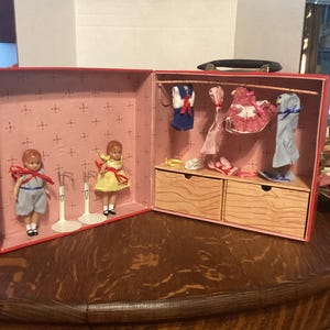 May include: A vintage dollhouse suitcase with two dolls and miniature clothing. The suitcase is red with a pink interior and features two dolls, one in blue and white striped overalls and the other in a yellow dress. The suitcase also contains miniature dresses and shoes.