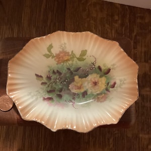 May include: A vintage, scalloped-edge dish with a floral design. The dish features a cream-colored interior with a peach-colored rim. The floral arrangement includes yellow and peach roses with green foliage. A penny is next to the dish.