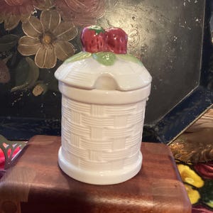 May include: White ceramic jar with a woven pattern and a lid shaped like a basket with two red strawberries on top.