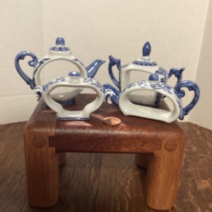 May include: Four miniature porcelain teapots and napkin rings with blue and white floral designs. The teapots are displayed on a small, dark wooden table. The teapots have curved handles and decorative lids. The napkin rings have a similar design.