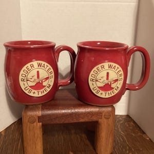 May include: Two red ceramic mugs with handles. Each mug features a circular emblem with the text "ROGER WATERS US+THEM" and a graphic of two hands. The mugs are displayed on a small wooden stand.