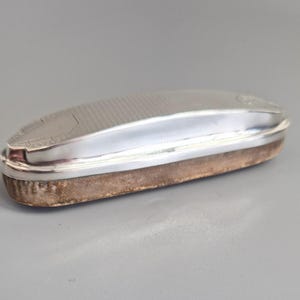 May include: A silver oval brush with a textured top and a brown suede-like base. The brush has a curved design and a polished, reflective surface. The item is likely an antique or vintage item.