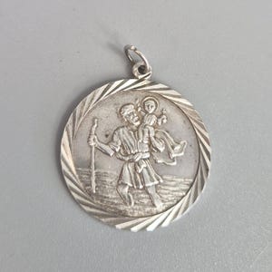 Może przedstawiać: Srebrny, okrągły wisiorek z szczegółowym reliefem przedstawiającym Świętego Krzysztofa niosącego dziecko. Wisiorek ma teksturowaną obwódkę i małą pętlę na łańcuszek. Projekt jest skomplikowany, a metal ma polerowane wykończenie.