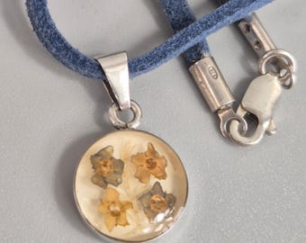 May include: A round pendant necklace with a silver-colored bezel. The pendant contains dried flowers in shades of yellow and brown, set against a cream background. The necklace has a blue suede cord with a silver-colored clasp.