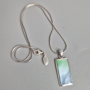 May include: A silver necklace with a rectangular pendant. The pendant features a gradient of green and blue. The chain is a thin, snake chain style, and the clasp is visible.