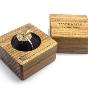 Puede incluir: Una caja de madera para anillo con un anillo de oro en su interior. La caja está grabada con las palabras "Para mi amada Margarita Te amo".