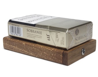 Puede incluir: Un paquete de cigarrillos Sobranie plateado con una etiqueta dorada y un código de barras en el lateral. El paquete está sobre un soporte de madera con un tornillo en la parte inferior.