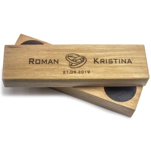 Puede incluir: Una caja de anillos de madera con un interior de terciopelo negro. La tapa está grabada con los nombres "Roman" y "Kristina", un dibujo de dos anillos entrelazados y la fecha "21.09.2019".